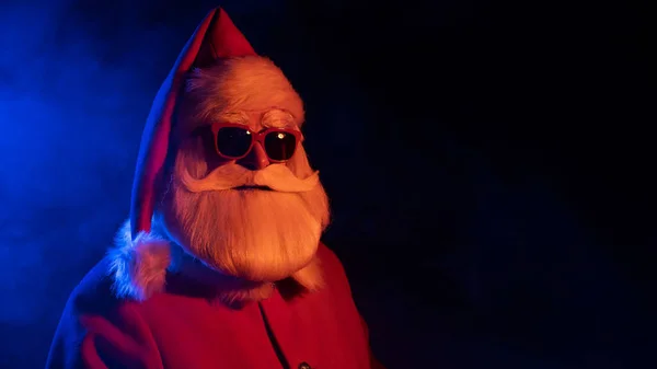 Święty Mikołaj w okularach przeciwsłonecznych w neonowym świetle na czarnym tle. Świąteczne przyjęcie. — Zdjęcie stockowe