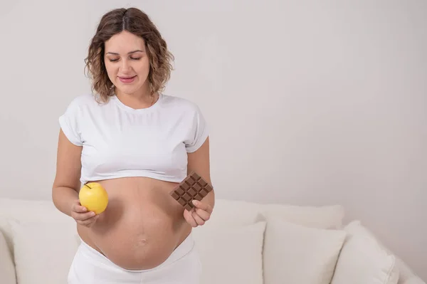 Беременная женщина держит яблоко и плитку шоколада. Девятый месяц беременности. — стоковое фото