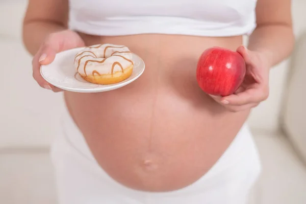 Беременная женщина держит красное яблоко и пончик. Выбор продуктов питания. — стоковое фото
