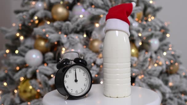 Una botella de leche con un sombrero de Santa Claus y un despertador en el fondo de un árbol de Navidad. Golosinas para Santa. — Vídeos de Stock
