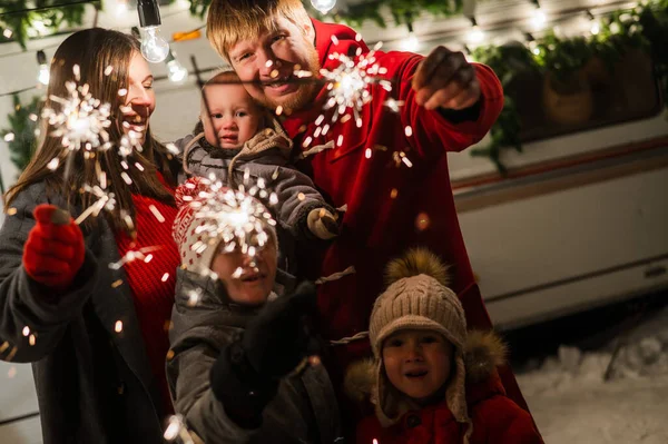Счастливая большая семья празднует Новый год на природе и держит в руках искры. Родители с тремя сыновьями путешествуют в передвижном доме. — стоковое фото