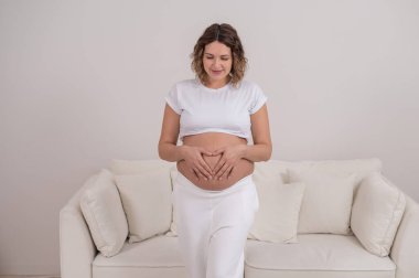 Hamile bir kadın ellerini karnında kalp şeklinde tutar..