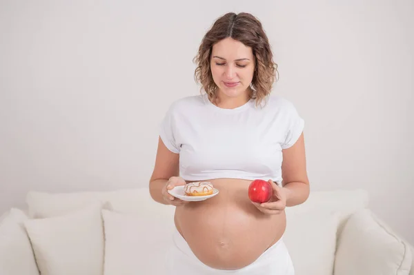 Egy terhes nő vörös almát és fánkot tart a kezében. Az élelmiszer kiválasztása. — Stock Fotó