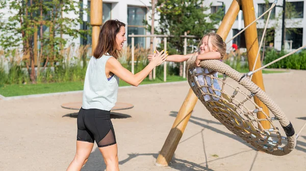 Mamma e figlia oscillano su un'altalena rotonda. Donna caucasica e bambina si divertono nel parco giochi. — Foto Stock