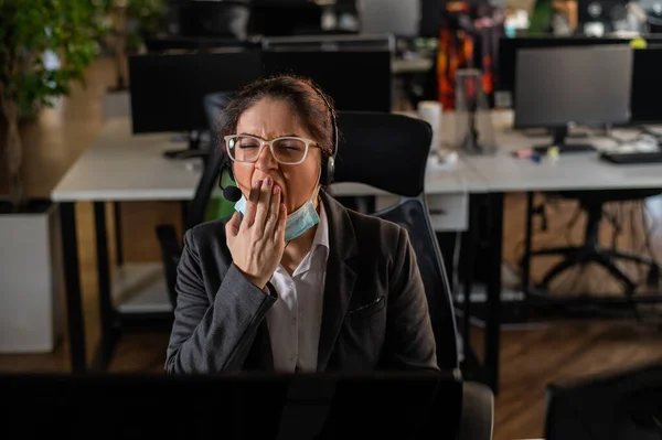 Affärskvinna i medicinsk mask gäspar vid sitt skrivbord på kontoret. — Stockfoto