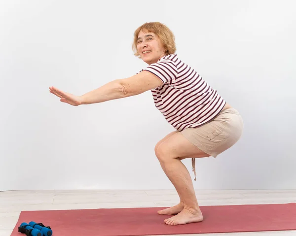 Äldre kvinna gör knäböj på en vit bakgrund. Den gamla damen gör övningar för sin hälsa — Stockfoto