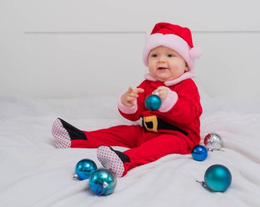 Noel Baba gibi giyinmiş bebek yatakta Noel süsleriyle oynuyor..