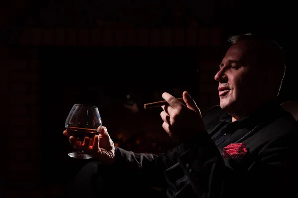 A mature man with glasses drinks brandy and smokes a cigar while sitting in an armchair by the fireplace in the dark. The concept of an elite gentlemens club