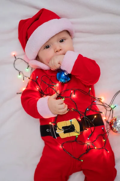 Chłopiec ubrany w strój Mikołaja leży na łóżku i bawi się świąteczną girlandą. — Zdjęcie stockowe