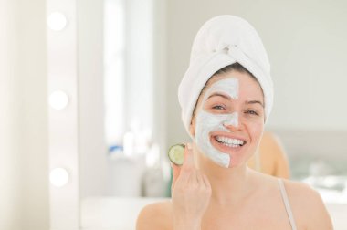 Saçında havluyla ve yüzünde kilden bir maskeyle elinde salatalıkla gezen neşeli bir kadın. Evdeki güzellikle ilgilenmek.