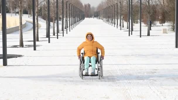 Běloška se zdravotním postižením jezdí v zimě na židli v parku. Dívka na procházce v invalidním vozíku. — Stock video