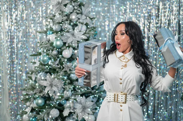 Sexy brunette in een witte jurk houdt geschenken door de kerstboom — Stockfoto