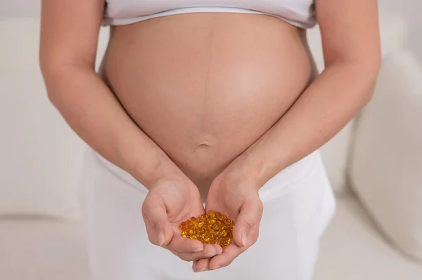 裸の腹を持つ妊婦はビタミンの一握りを保持します。透明な黄金の丸薬。期待する母親のためのビタミンD. — ストック写真