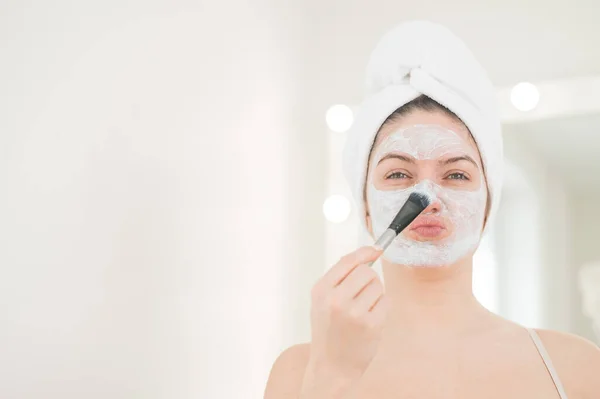 Hermosa mujer caucásica con una toalla en el pelo se aplica una mascarilla de barro. Cuidar de la belleza en casa — Foto de Stock