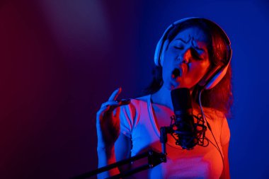 Gözlüklü ve kulaklıklı beyaz kadın siyah arka planda neon ışıkta mikrofona şarkı söylüyor. Duygusal bir kız kayıt stüdyosunda şarkı kaydediyor.