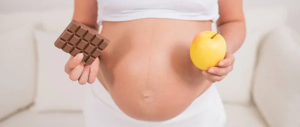 Uma grávida está segurando uma maçã e uma barra de chocolate. O nono mês de gravidez. — Fotografia de Stock