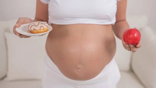 Uma mulher grávida está segurando uma maçã vermelha e um donut. Escolha de alimentos. — Fotografia de Stock