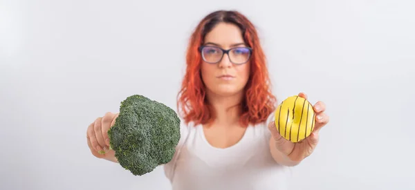 Vit kvinna väljer mellan grönsaker och snabbmat. rödhårig flicka som håller broccoli och munk på en vit bakgrund. — Stockfoto