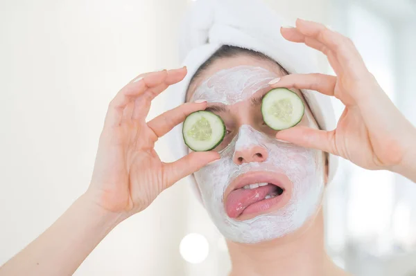 Cheerful woman with a towel on her hair and in a clay face mask fooling around with cucumbers in her hands. Taking care of beauty at home — Stock Photo, Image