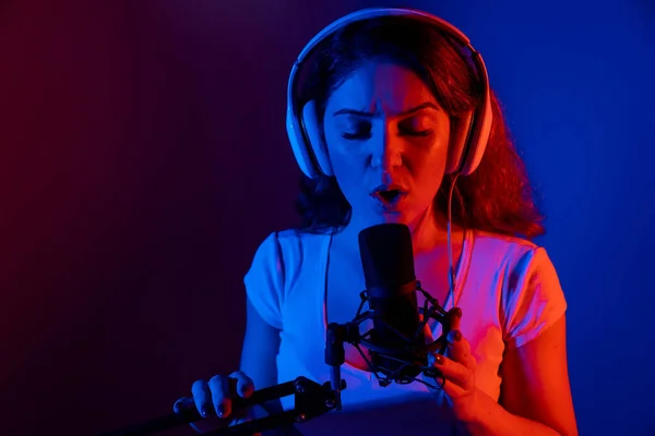 Fehér nő szemüvegben és fülhallgatóban neon fényben énekel egy mikrofonba, fekete háttérrel. Egy érzelmes lány felvesz egy dalt egy stúdióban. — Stock Fotó