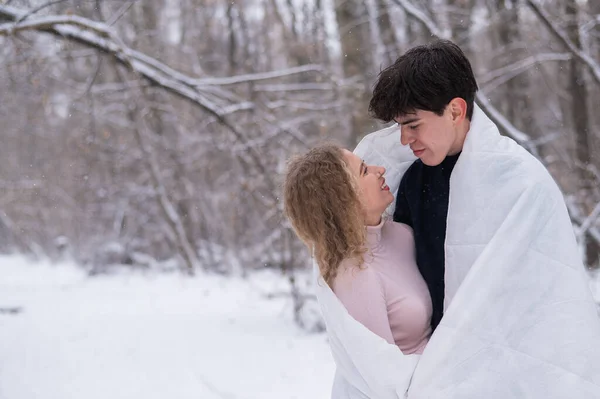 Un jeune couple se promène dans le parc en hiver. Le mec et la fille s'embrassent enveloppés dans une couverture blanche à l'extérieur. — Photo