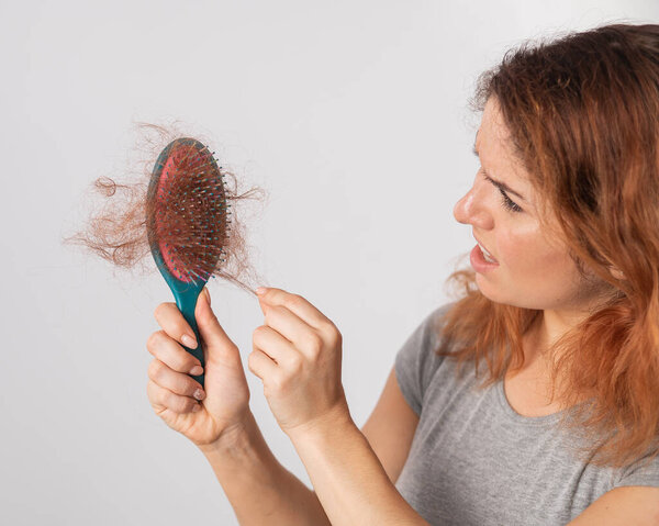 Белая женщина с гримасой ужаса держит расческу с булочкой волос. Выпадение волос и женская лысина.
