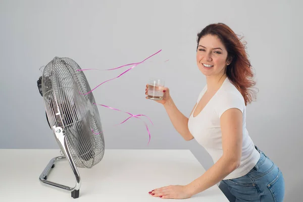 En rödhårig vit kvinna ryser av den elektriska fläkten och dricker en kall drink. Klimatkontroll i lägenheten — Stockfoto