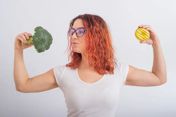 Vit kvinna väljer mellan grönsaker och snabbmat. rödhårig flicka som håller broccoli och munk på en vit bakgrund. — Stockfoto