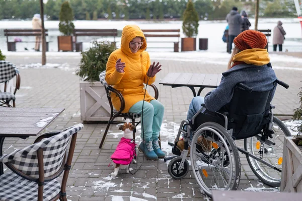 Two girlfriends in a cafe on a street terrace in winter. Woman in a wheelchair. — Stockfoto