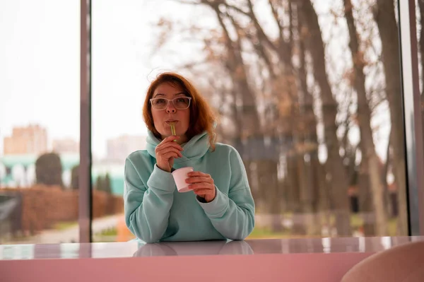 Caucásica pelirroja mujer come helado con una cuchara de plástico mientras está sentada sola en un café. —  Fotos de Stock