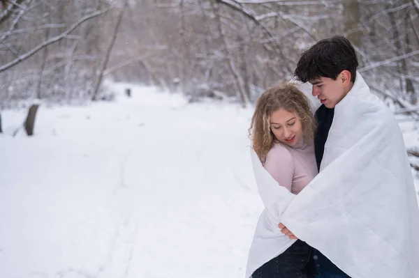 Um jovem casal caminha no parque no inverno. O cara e a menina estão se beijando envolto em um cobertor branco ao ar livre. — Fotografia de Stock
