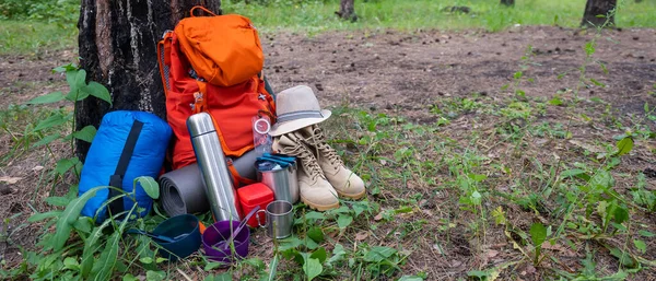 Çam ormanında yürüyüş ekipmanları. Sırt çantası, termos, uyku tulumu, pusula, şapka ve ayakkabılar. — Stok fotoğraf