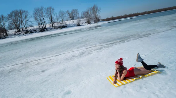 Donna caucasica in costume da bagno rosso e cappello a maglia prende il sole in inverno sdraiata sulla neve. — Foto Stock