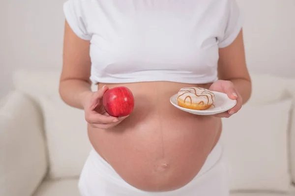 Egy terhes nő vörös almát és fánkot tart a kezében. Az élelmiszer kiválasztása. — Stock Fotó