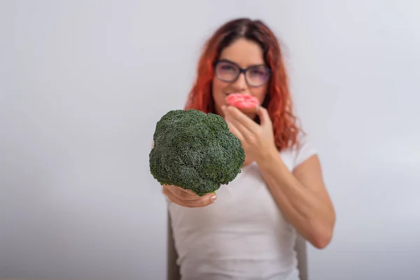 Vit kvinna väljer mellan grönsaker och snabbmat. rödhårig flicka äta pochik och hålla broccoli på en vit bakgrund — Stockfoto