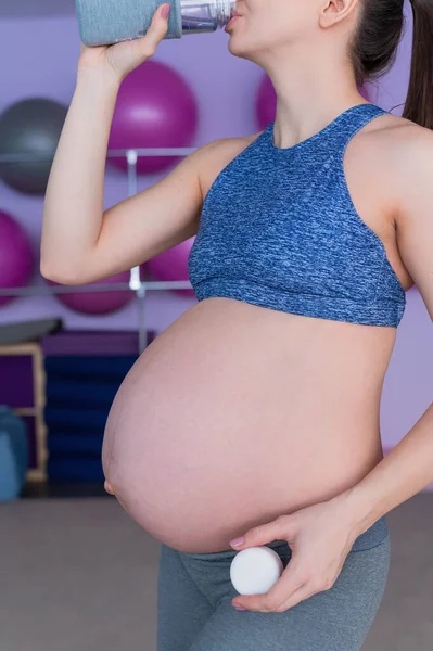 Une femme enceinte en tenue de sport boit de l'eau dans le gymnase avec des épées de fitness. La fille est engagée à Pilates pour les femmes enceintes. Dernier trimestre . — Photo
