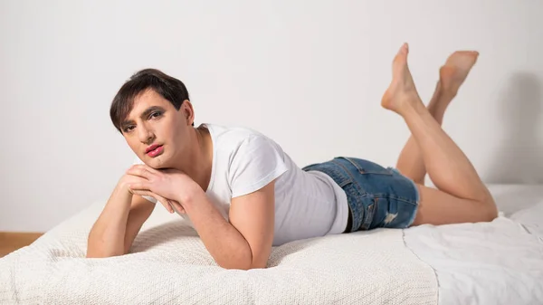 L'uomo transgender caucasico giace sul letto gay. all'aperto gay posa in il camera da letto. — Foto Stock