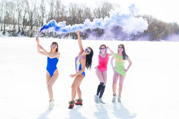 Quatro mulheres caucasianas de fato de banho têm bombas de fumo coloridas numa praia nevada. Meninas quentes posando em biquínis ao ar livre no inverno. — Fotografia de Stock