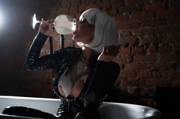 Mulher sexy em peruca branca curta e terno de látex bebe leite de um copo enquanto está sentado no banheiro. — Fotografia de Stock