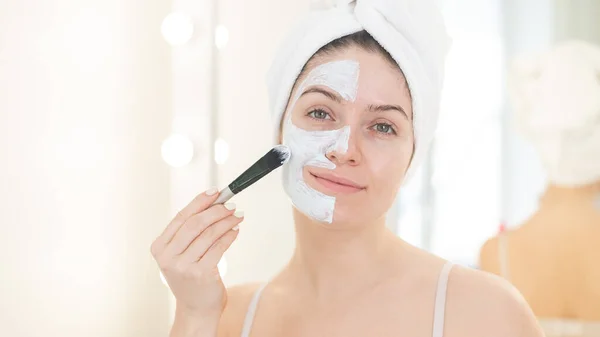 Piękna biała kobieta z ręcznikiem na włosach nakłada glinianą maskę na twarz. Dbanie o piękno w domu — Zdjęcie stockowe