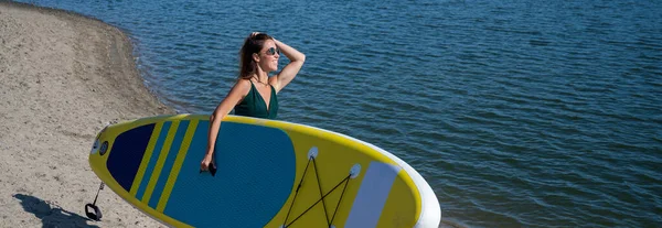 Kavkazská žena kráčí po pláži a nese sup board na řece ve městě. Letní sport. — Stock fotografie
