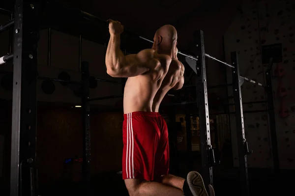 Blanke man met een naakte romp trekt op de horizontale balk in de sportschool. — Stockfoto