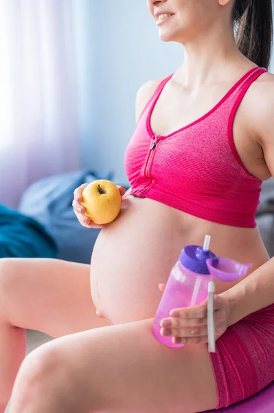 Yogadan sonra elma yiyen yüzü olmayan hamile kadın. Müstakbel anne pilates yaptıktan sonra meyve yer ve su içer. Fitness topunda oturan kız. — Stok fotoğraf