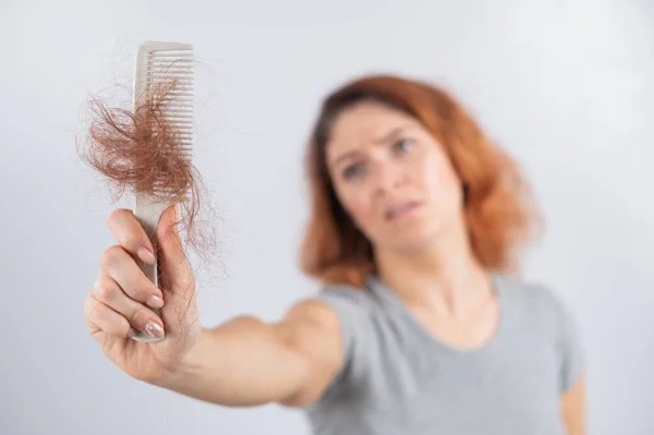 Caucasian woman with a grimace of horror holds a comb with a bun of hair. Hair loss and female alopecia. — Stock Photo, Image