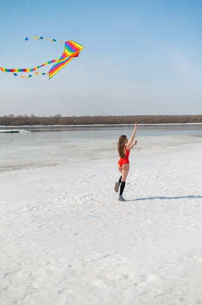 Caucasian woman in a red swimsuit launches a kite on the seashore in winter. — Stock Photo, Image