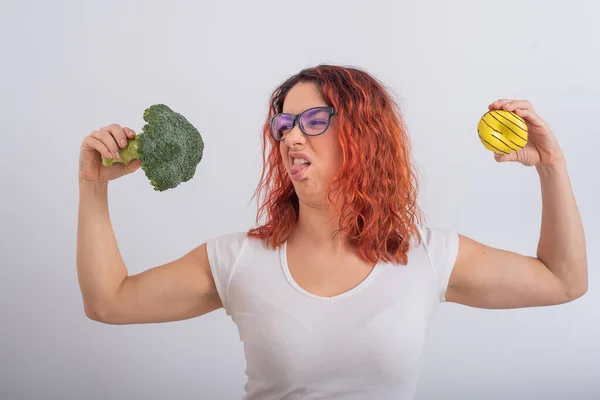 Vit kvinna väljer mellan grönsaker och snabbmat. rödhårig flicka som håller broccoli och munk på en vit bakgrund. — Stockfoto