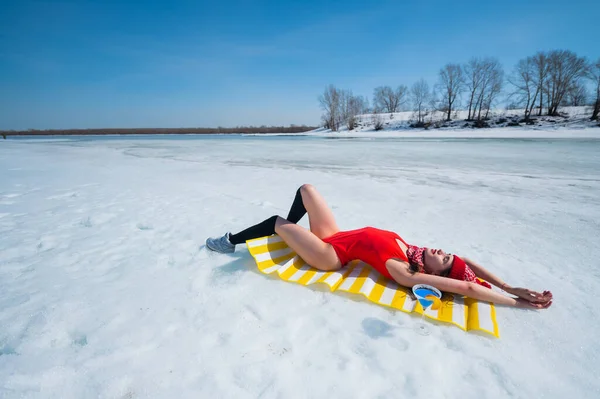 Donna caucasica in costume da bagno rosso e cappello a maglia prende il sole in inverno sdraiata sulla neve. — Foto Stock