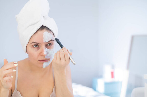 Beautiful caucasian woman with a towel on her hair applies a clay face mask. Taking care of beauty at home