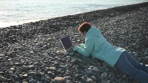 Mujer caucásica acostada boca abajo en una playa de guijarros y escribiendo en un portátil. Trabajo remoto freelancer. — Vídeos de Stock