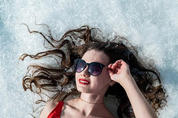 Close-up portrait of a caucasian woman lying on the snow in sunglasses. Top view. — Stock Photo, Image